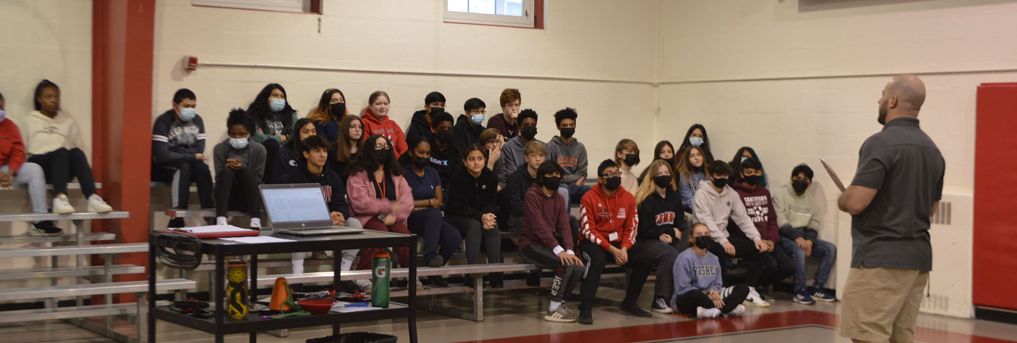 Students sit in bleachers and listen to teacher in gymnasium
